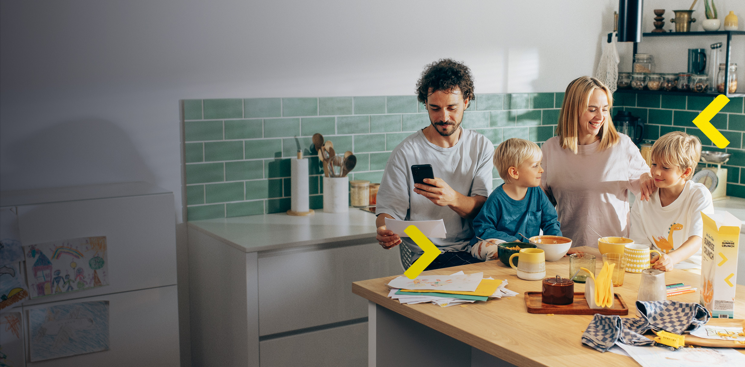 Család a konyhában reggelizik, az apa mobilon intézi a pénzügyeit.